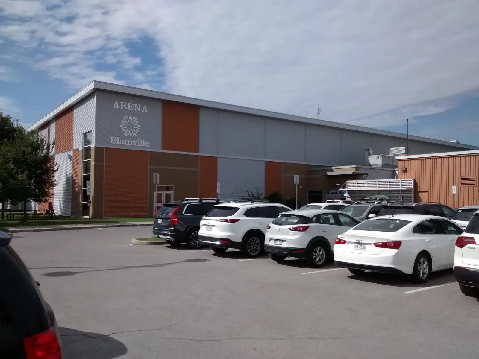 La façade orange et grise de l’aréna, avec l’enseigne ARÉNA Blainville au-dessus de l’entrée, vue de l’autre côté du stationnement. — Aréna de Blainville