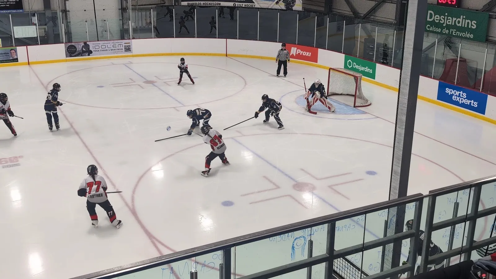 Un match vu d’au-dessus de la baie vitrée, des joueurs répartis autour d’un filet au bout d’une grande surface de glace, avec des panneaux de commanditaires le long du mur. — Aréna de Blainville