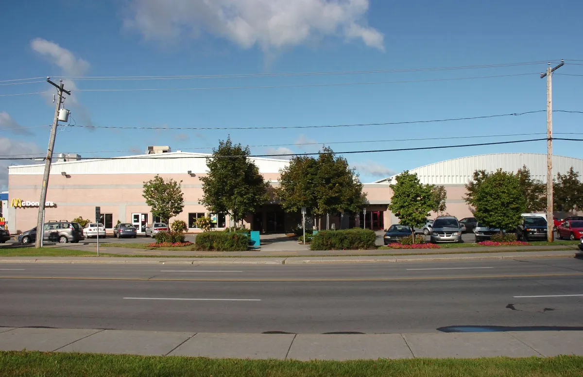 La larga arena rosa y beige con su techo curvo blanco detrás, vista desde el otro lado del bulevar con autos estacionados al frente. — Aréna de Boisbriand