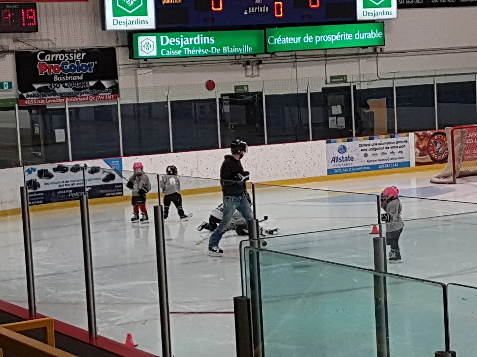 Patinadores repartidos por la pista vistos por encima de un barandal verde y rojo, con un marcador y un letrero de Desjardins sobre las vallas del fondo. — Aréna de Boisbriand