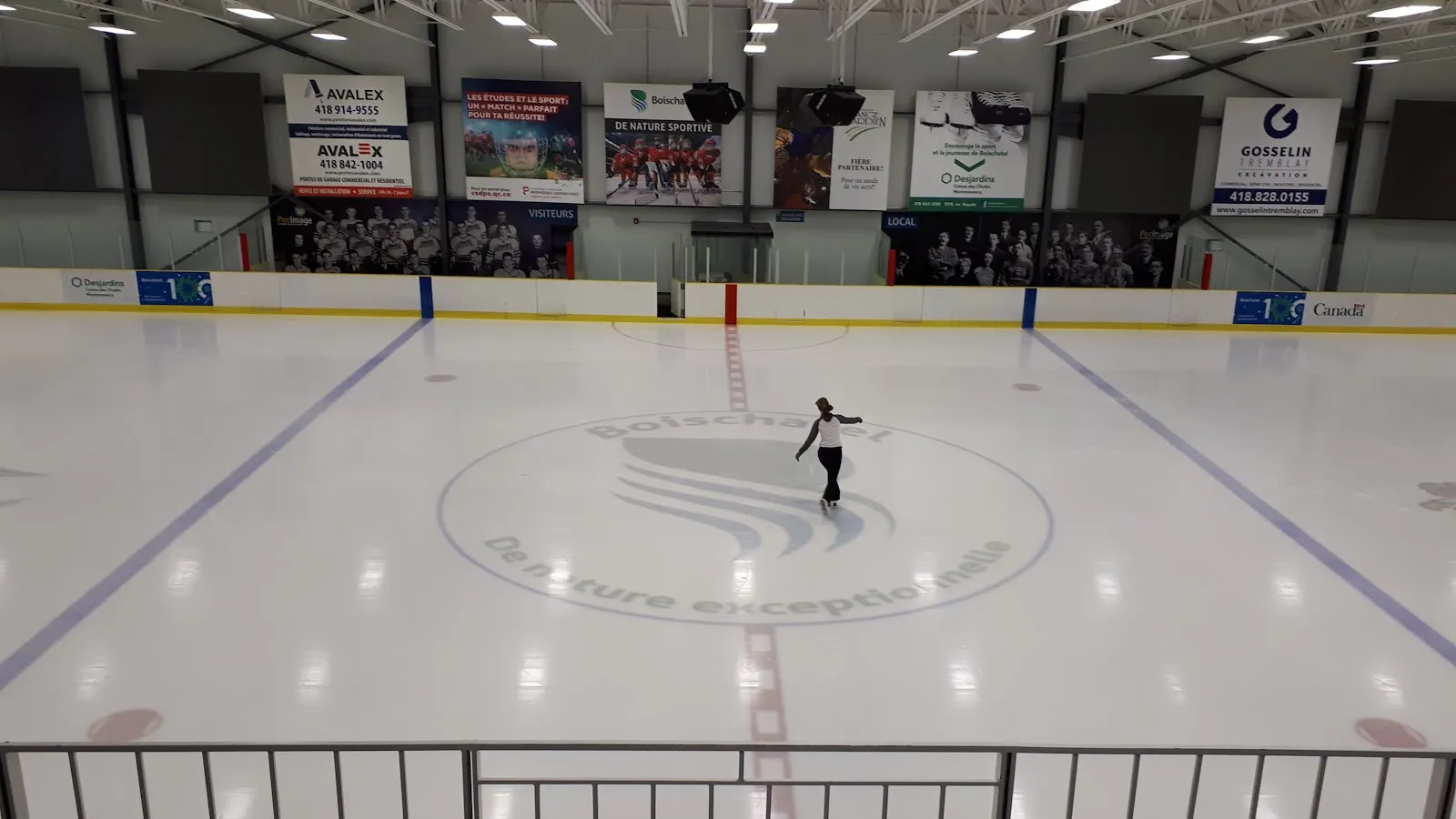 La surface de glace de l’Aréna de Boischatel, avec le logo du club au centre, un patineur seul et les bandes de commanditaires en arrière-plan. — Aréna de Boischatel