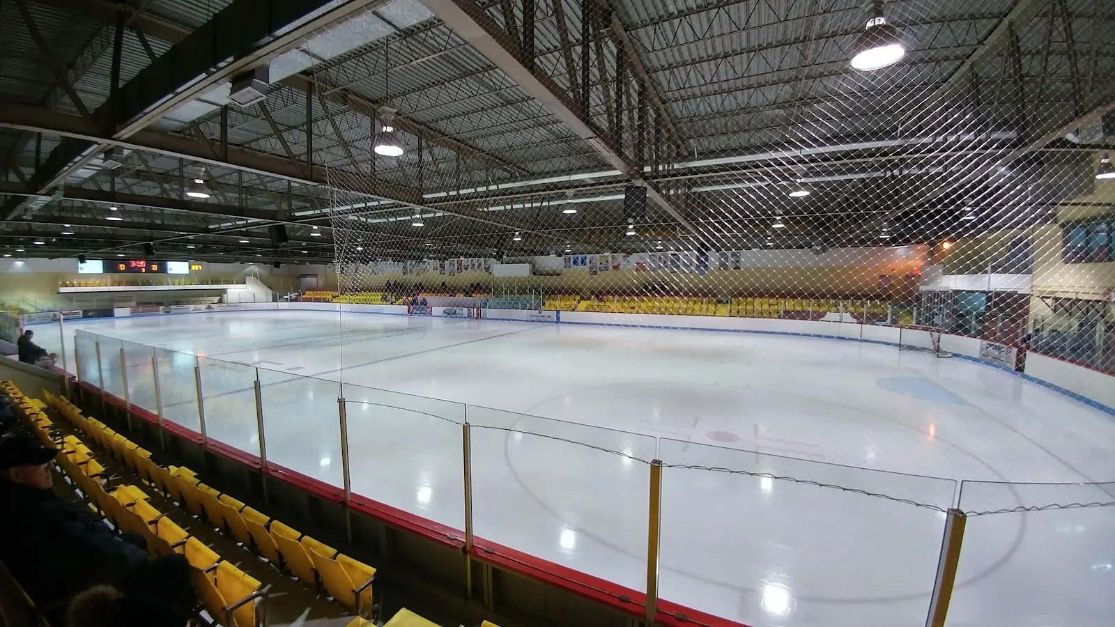 Une vue large de la glace depuis les gradins, une glace blanche vide à l’intérieur d’une rampe courbe, avec des bancs jaunes du côté proche. — Aréna de Clermont