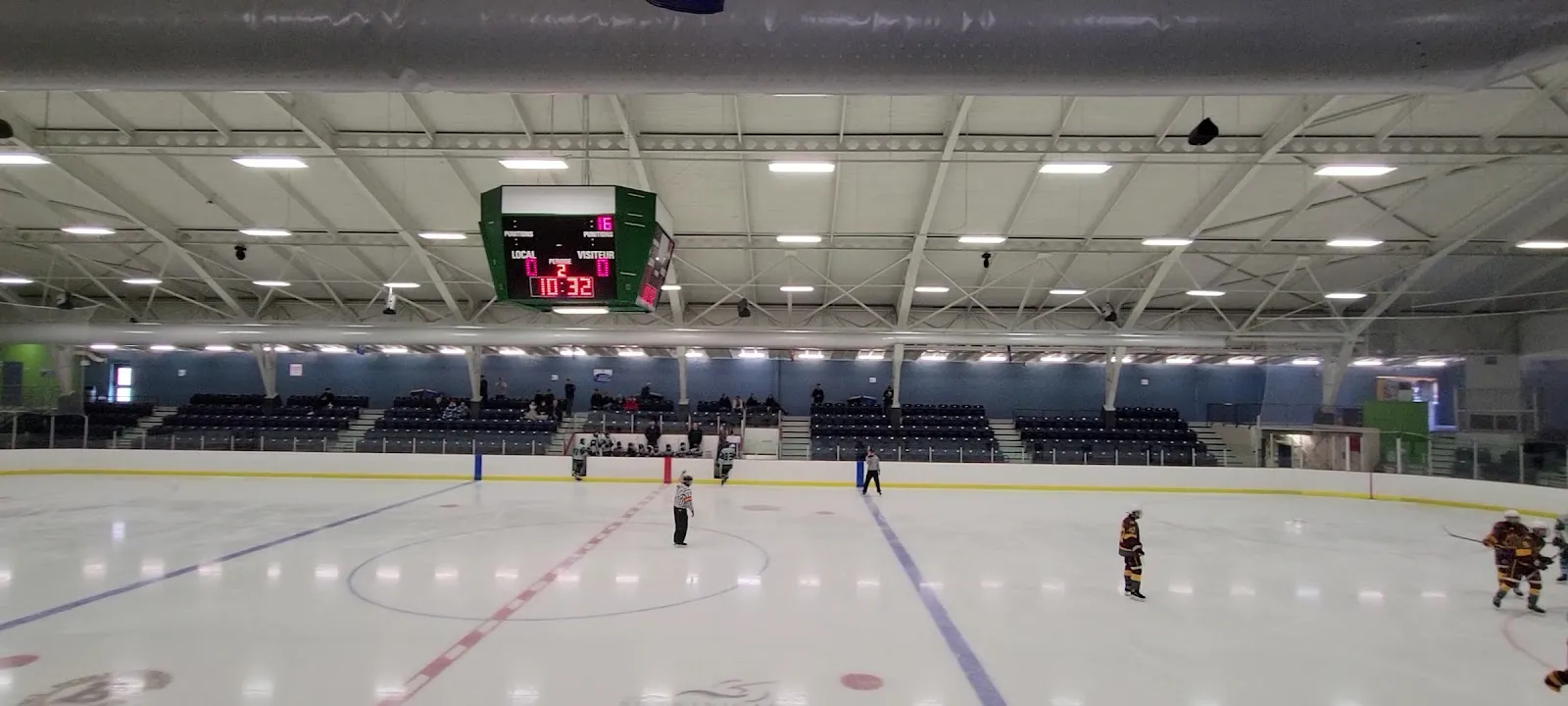 The ice of the Aréna de Grand-Mère seen from the stands, with the scoreboard hanging over centre ice and the steel roof trusses — Aréna de Grand-Mère