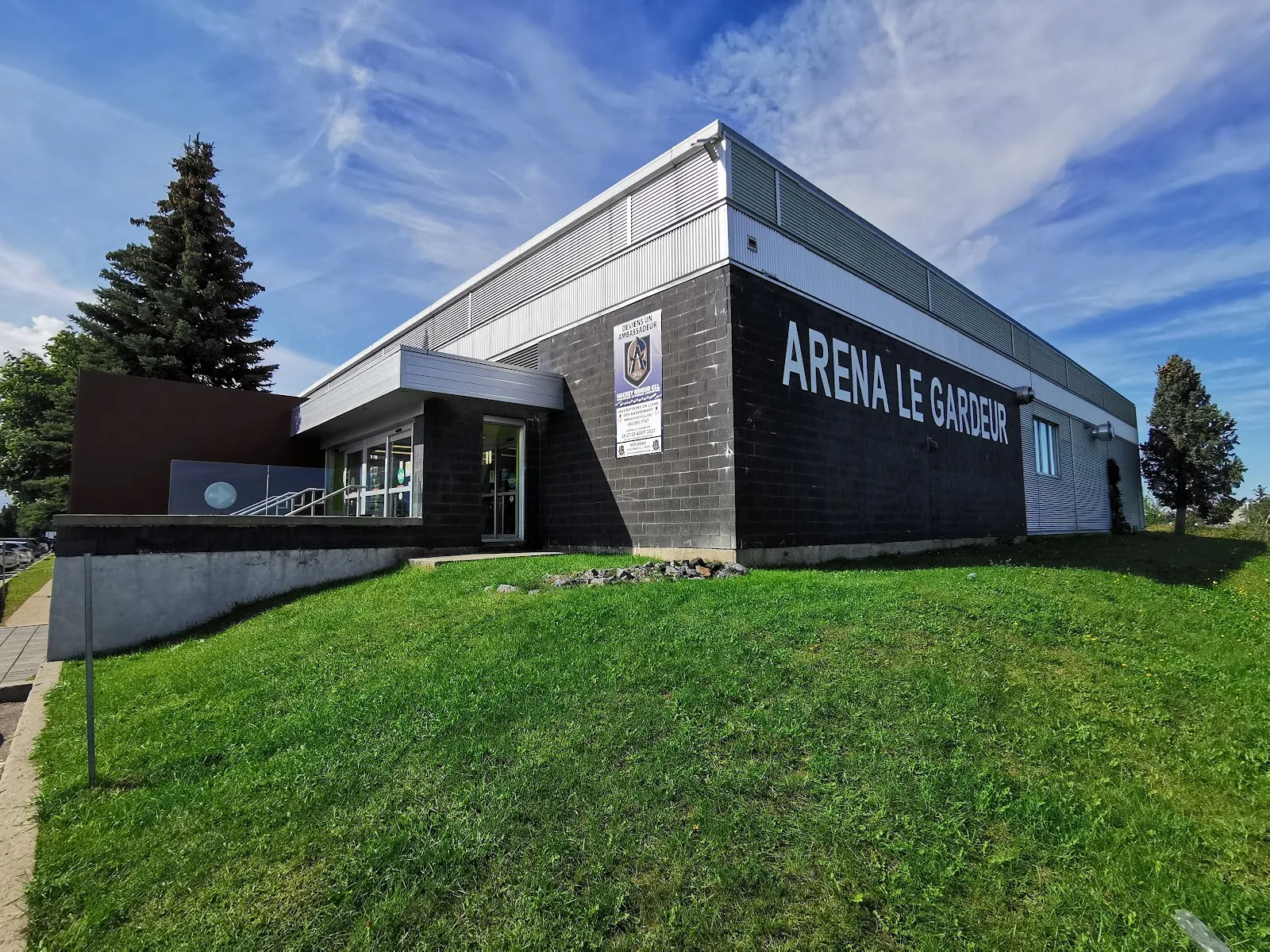 El Aréna Le Gardeur visto desde el césped en un día luminoso: un edificio bajo de revestimiento oscuro con el nombre Arena Le Gardeur en la fachada y un abeto al lado. — Aréna de Le Gardeur