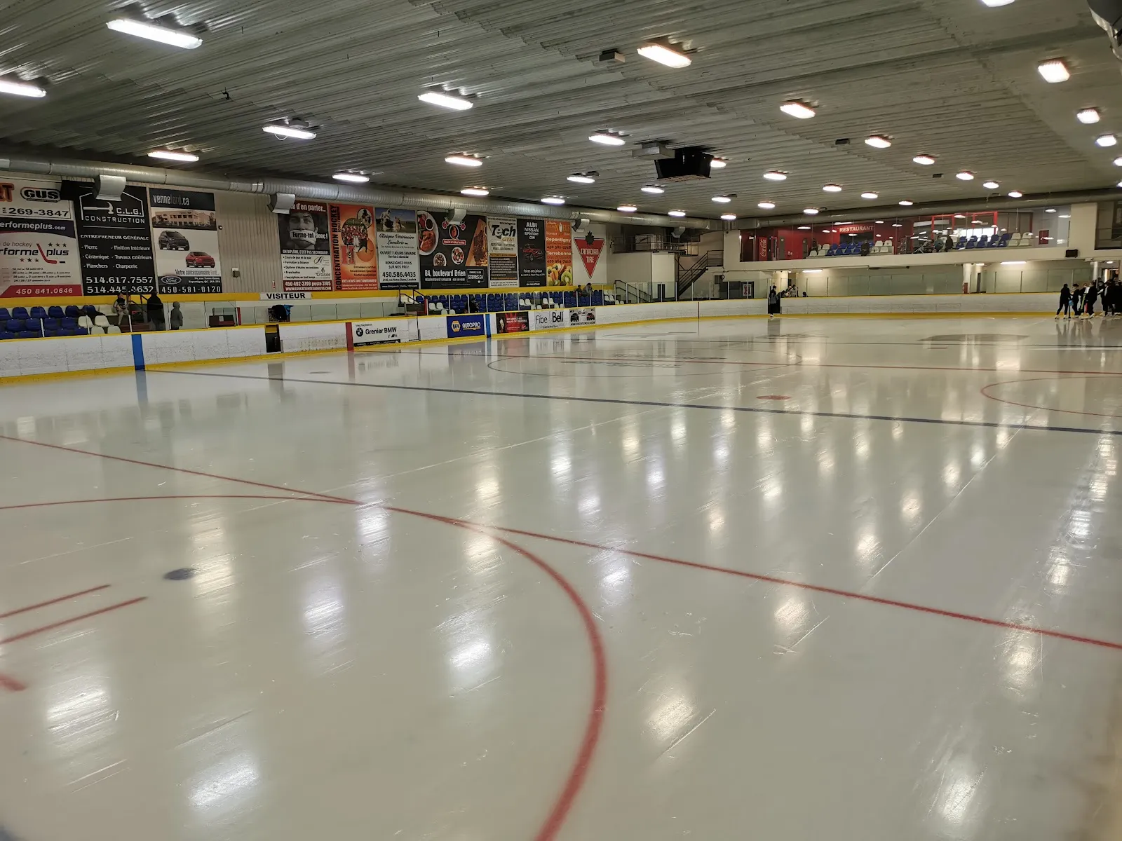 La pista vacía y recién renovada al nivel del hielo, con anuncios de patrocinadores a lo largo de la pared del fondo y un techo blanco bajo con hileras de luces. — Aréna de Le Gardeur