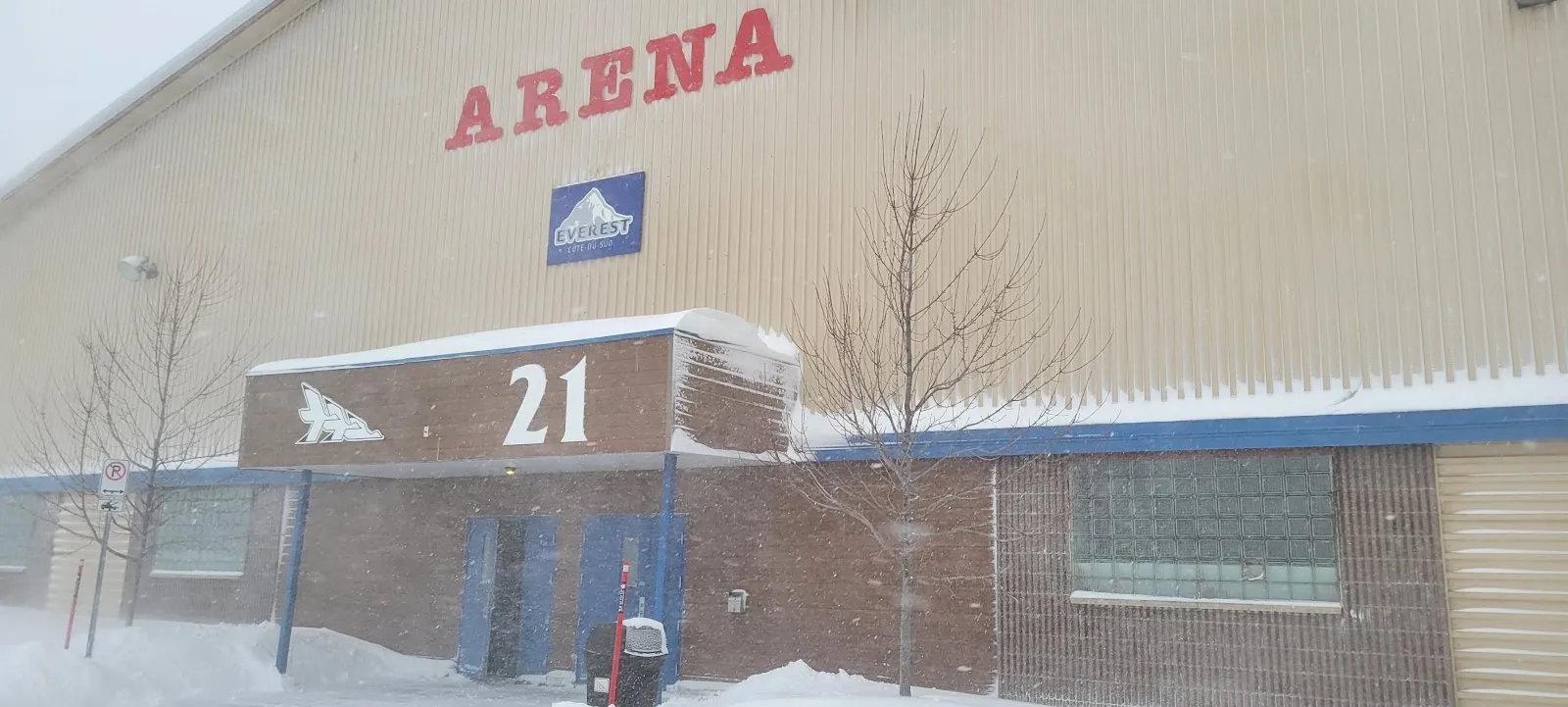 La esquina del Aréna de Montmagny con la palabra ARENA en mayúsculas rojas sobre una entrada azul, con nieve en el suelo y un árbol sin hojas junto a la pared. — Aréna de Montmagny