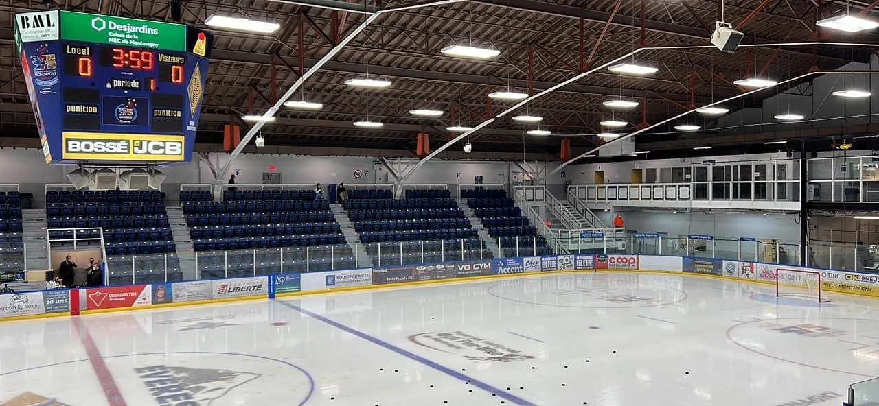 Toda la pista desde una esquina, con asientos azules a ambos lados y el marcador sobre el hielo. — Aréna de Montmagny