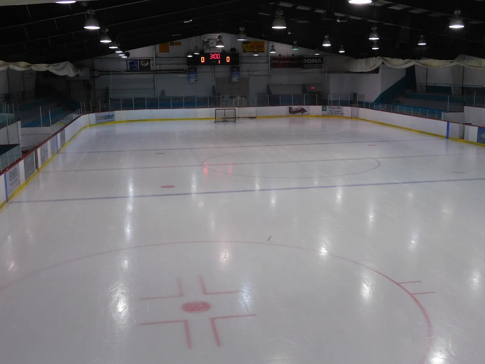 The empty ice sheet of the New Richmond arena with its red and blue lines, the scoreboard above the far boards and teal seating along the side. — Aréna de New Richmond