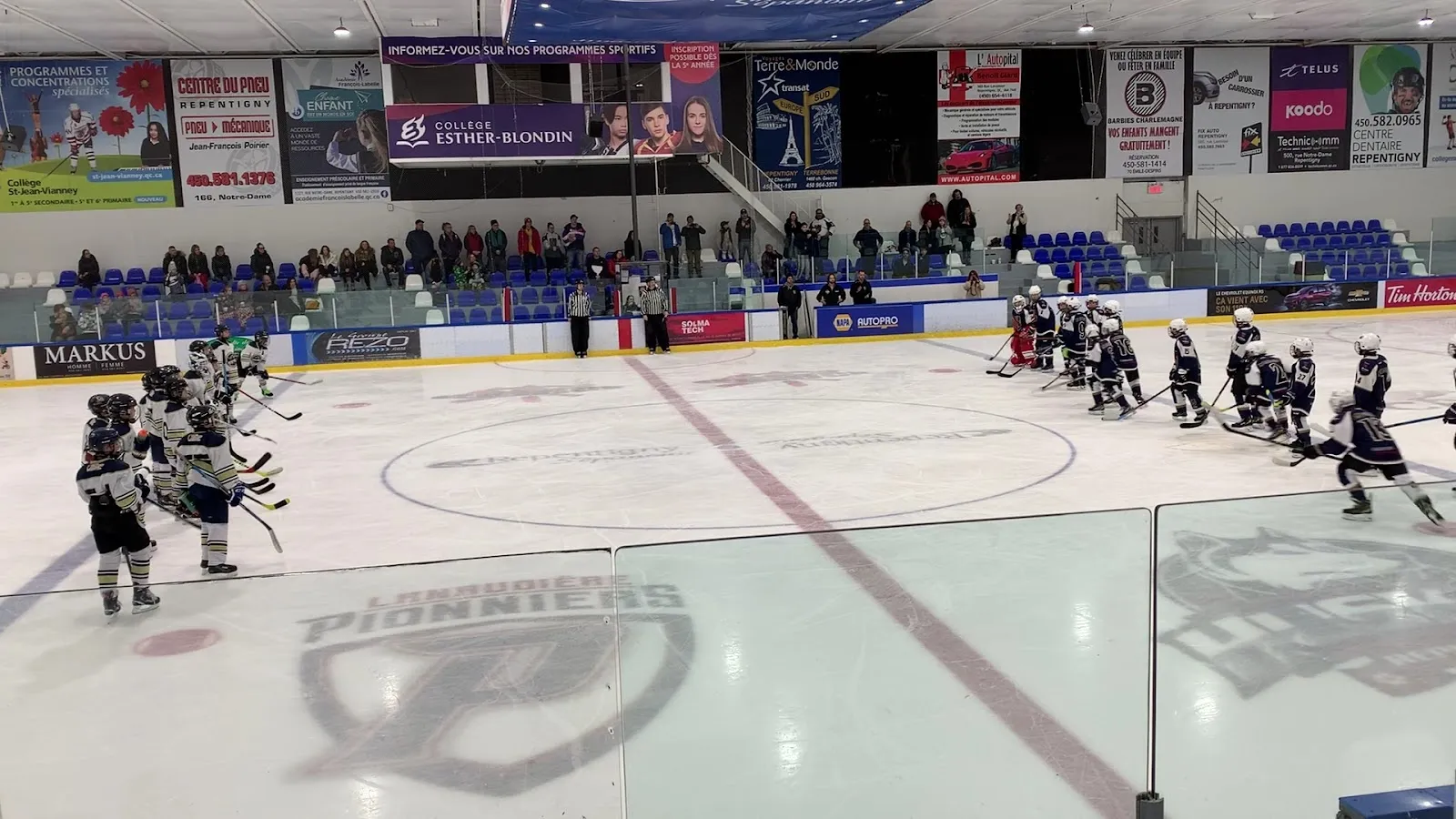 Un partido en el extremo cercano de una pista amplia bajo una pared con banderines, con jugadores repartidos alrededor de un logotipo en el centro y espectadores en el lado opuesto. — Aréna de Repentigny