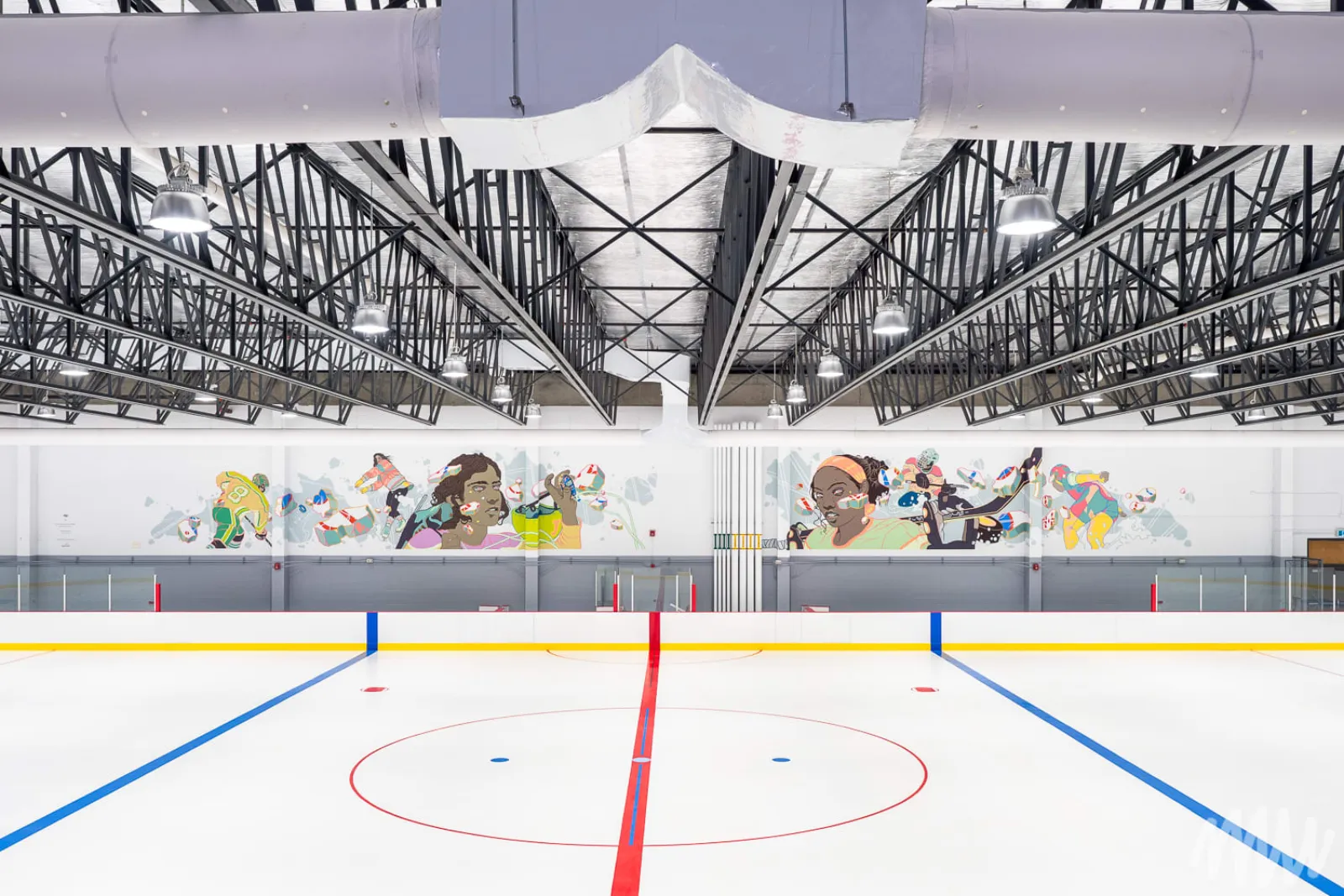 La patinoire vide avec la ligne rouge, les lignes bleues et le cercle central; des murales sportives colorées sur le mur du fond, et des fermes d’acier et des conduits apparents au plafond. — Aréna de Saint-Michel