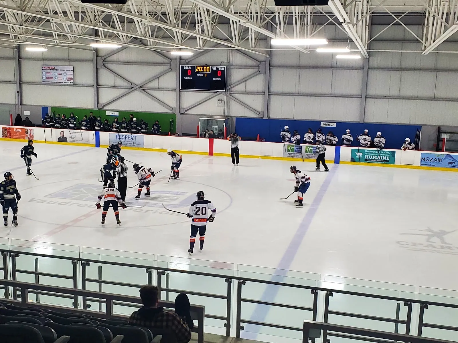 Un partido visto a través de la pista bajo un techo de acero blanco, con jugadores repartidos por el hielo y vallas de patrocinadores verdes y azules en el lado opuesto. — Aréna de Vaudreuil-Dorion
