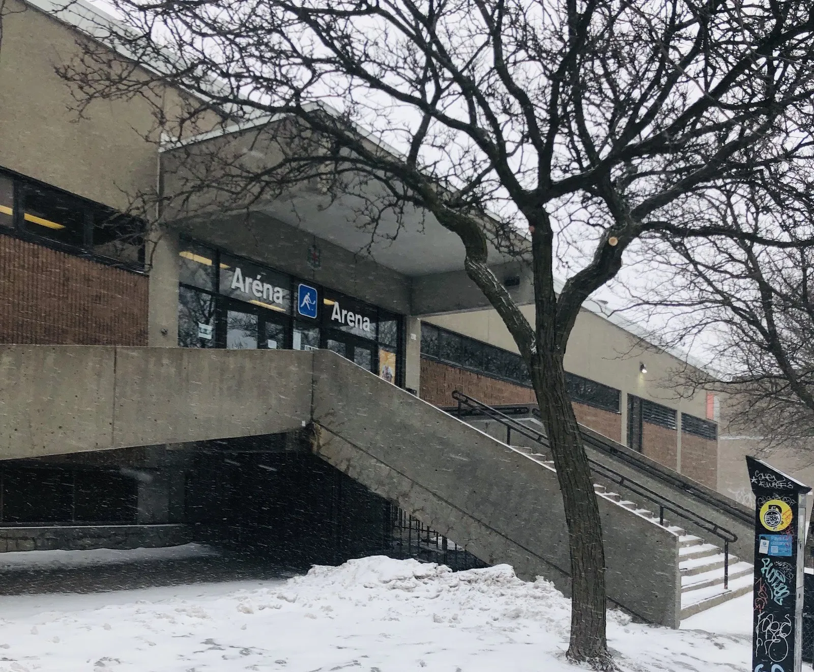 L’entrée de l’Aréna des Patineuses sous la neige : l’enseigne bilingue « Aréna/Arena » au-dessus des portes vitrées, auxquelles mène un escalier de béton — Aréna des Patineuses