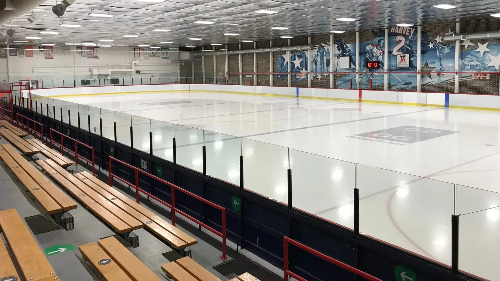 Empty ice rink seen from the stands, with a wall mural honoring Doug Harvey (retired number 2) and other hockey figures at the far end, red-railed wooden bleachers in the foreground — Aréna Doug-Harvey