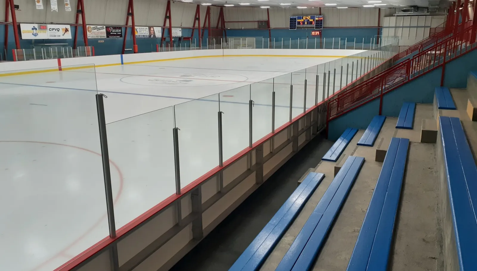 The ice seen from the blue bench bleachers, with red trim along the boards and red roof trusses above. — Aréna Duberger