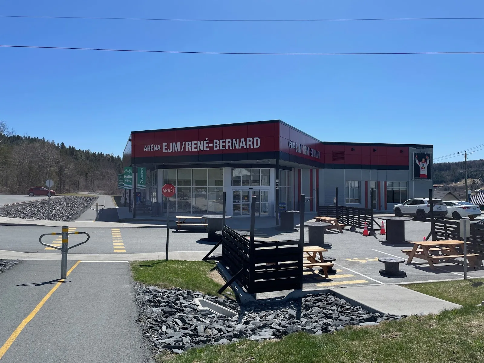 The arena's glass-fronted entrance with red 'ARÉNA EJM/RENÉ-BERNARD' signage across the top, picnic tables on a paved terrace and the bike path in the foreground, under a clear blue sky. — Aréna EJM/René-Bernard