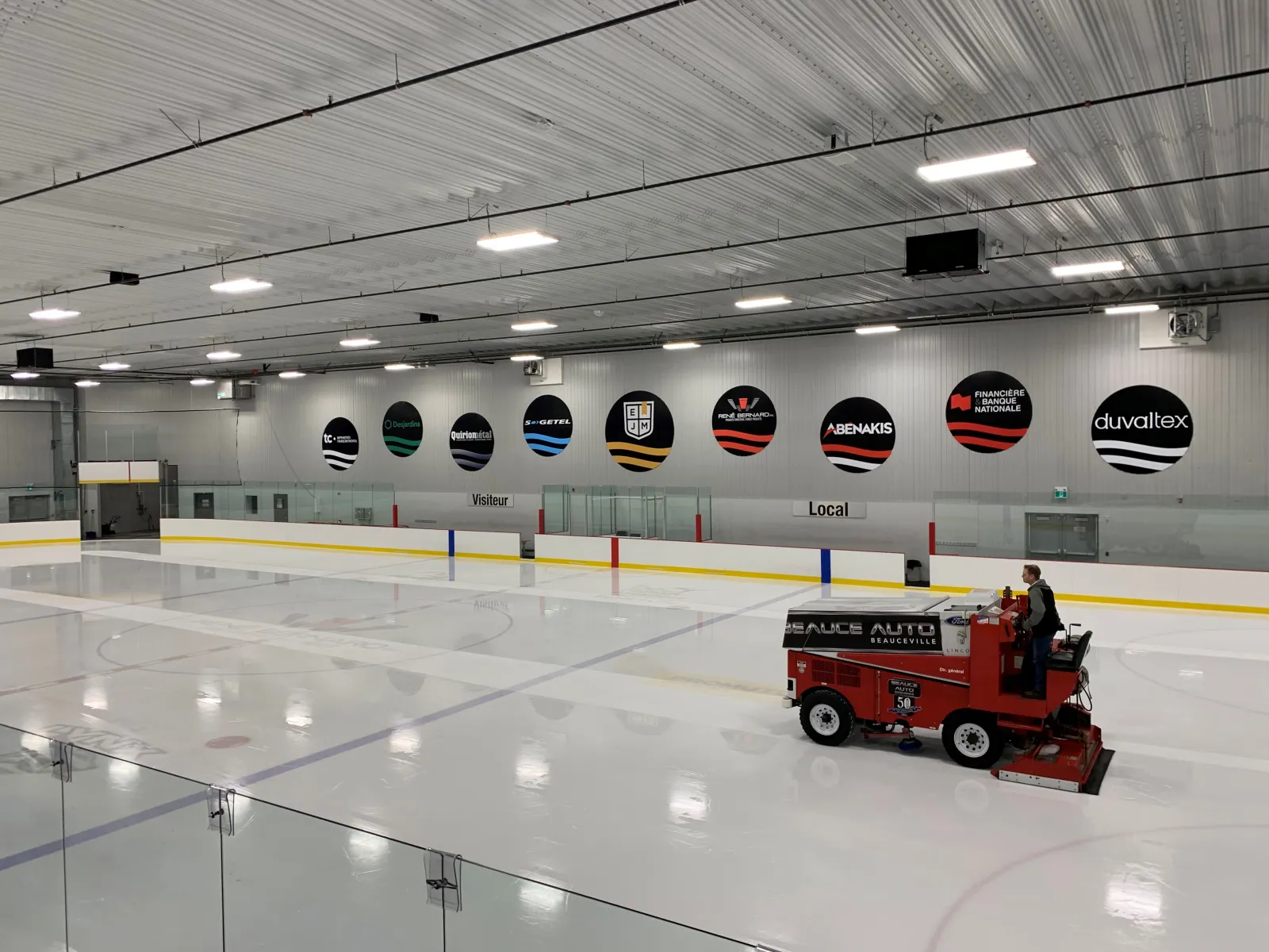 The empty ice sheet seen from rinkside glass, with the ice resurfacer at work, a row of round sponsor logos on the far wall and a white steel roof. — Aréna EJM/René-Bernard