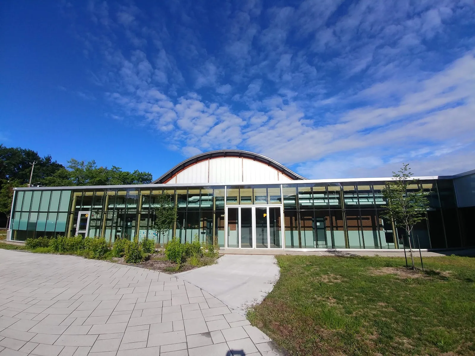La façade vitrée de l’Aréna Eric-Sharp et son toit courbé, derrière un sentier pavé et une pelouse. — Aréna Eric-Sharp