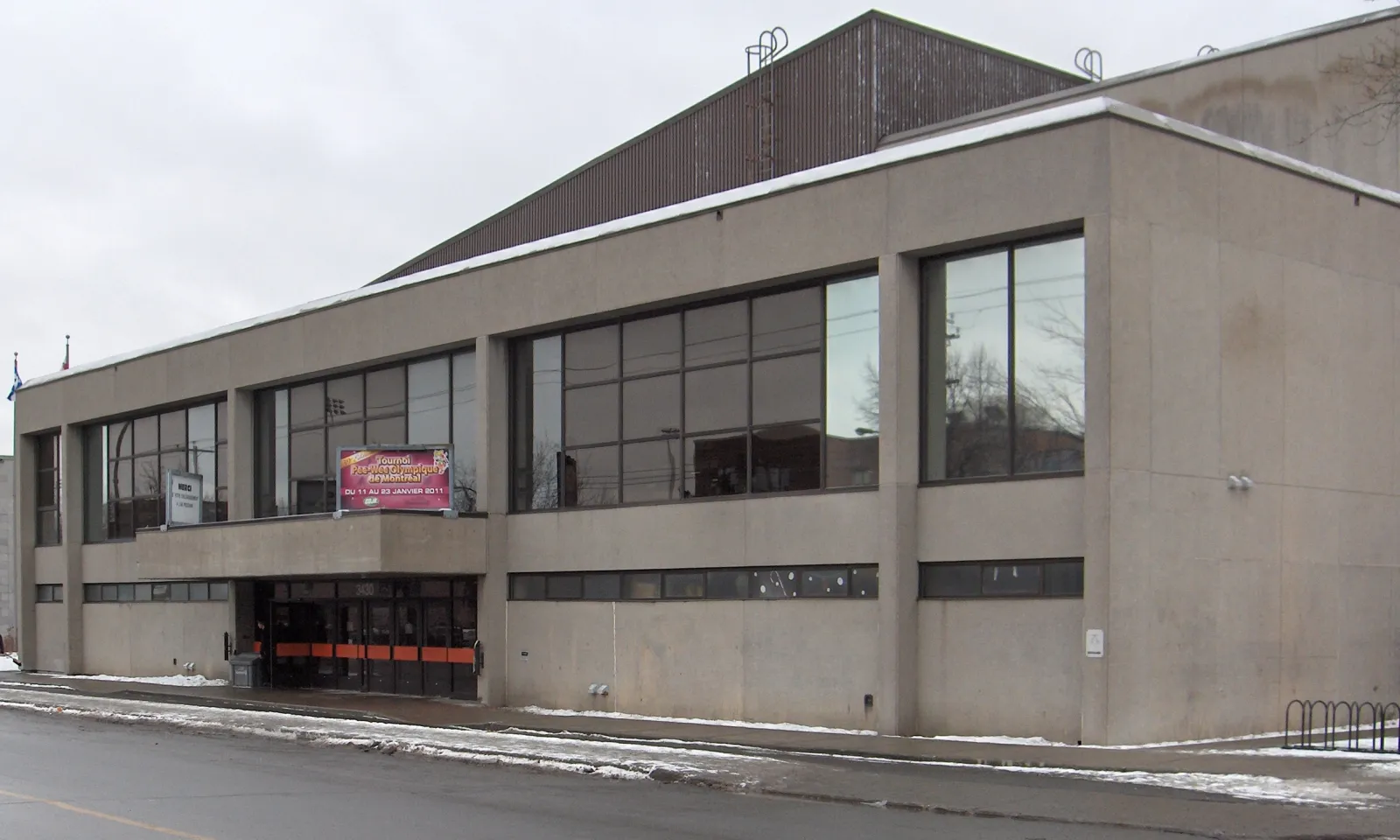 Vue au niveau de la rue de l’entrée principale de l’aréna en béton, avec le numéro 3430 au-dessus des portes et les drapeaux du Québec et du Canada sur des mâts près de la façade. — Aréna Étienne-Desmarteau
