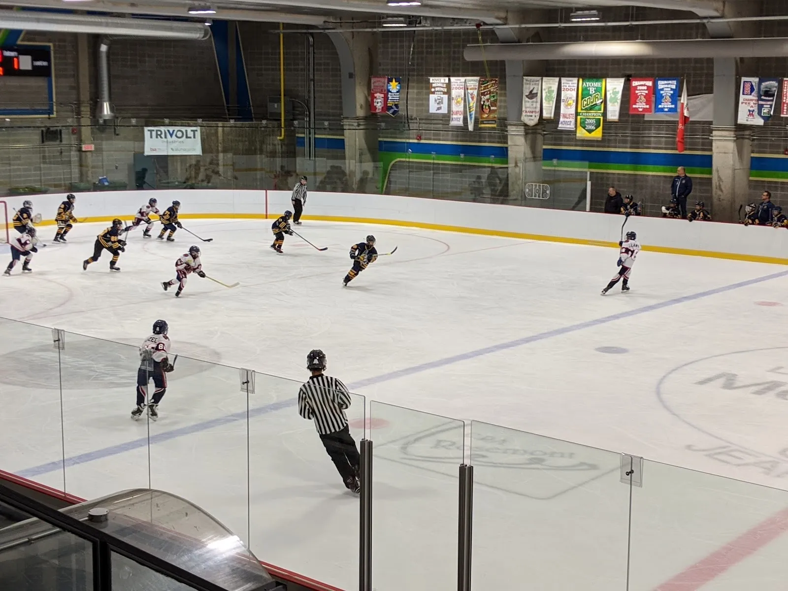 Un match vu par-dessus la baie vitrée sur une grande surface de glace, des joueurs répartis près des bandes du fond, avec des bannières de couleur accrochées au mur derrière. — Aréna Étienne-Desmarteau