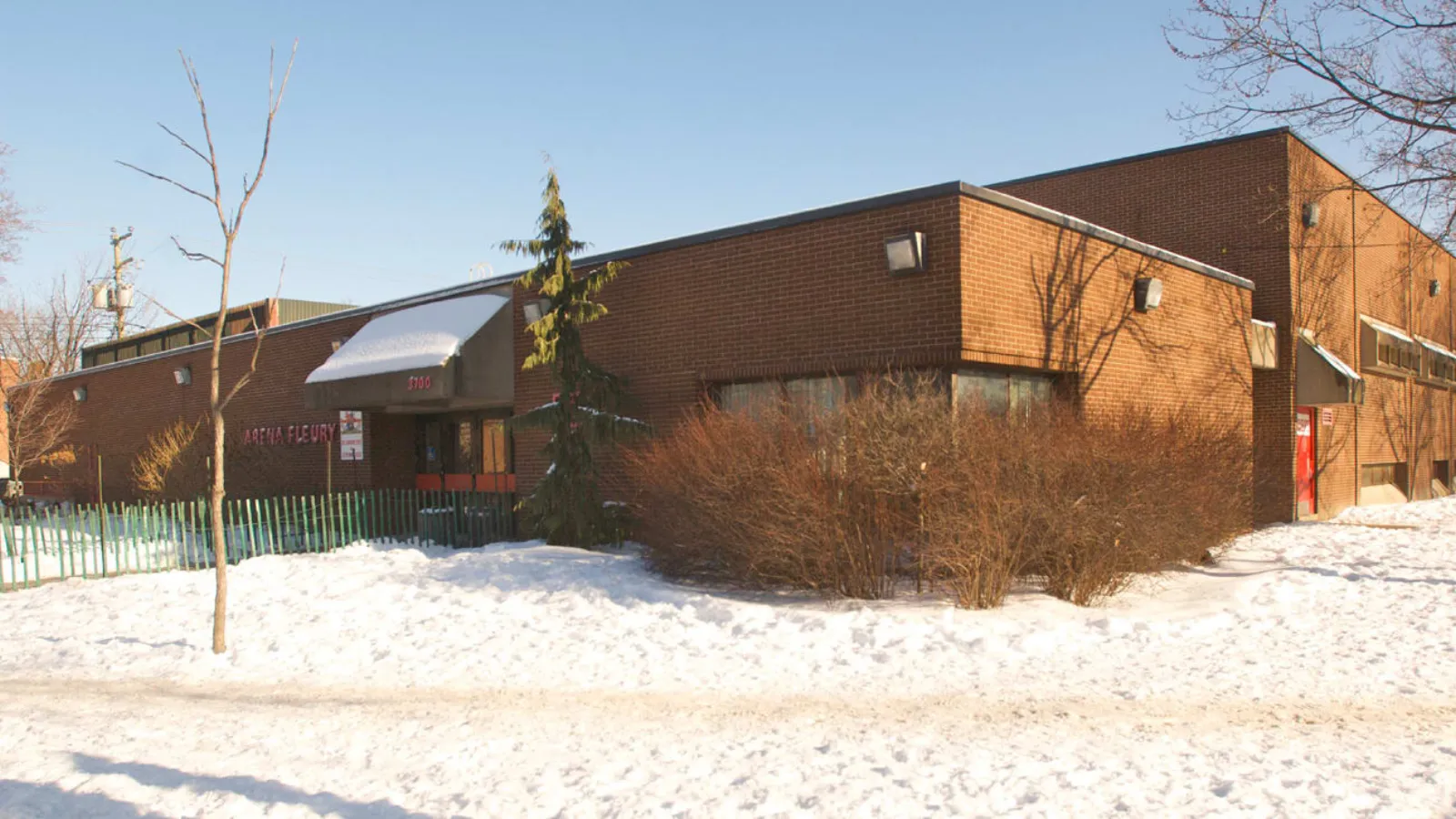 Vue hivernale enneigée d’un bâtiment d’aréna d’un étage en brique, avec une enseigne rouge « ARENA FLEURY » et un auvent avec le numéro civique au-dessus de l’entrée, des arbustes dénudés et un petit conifère devant. — Aréna Fleury
