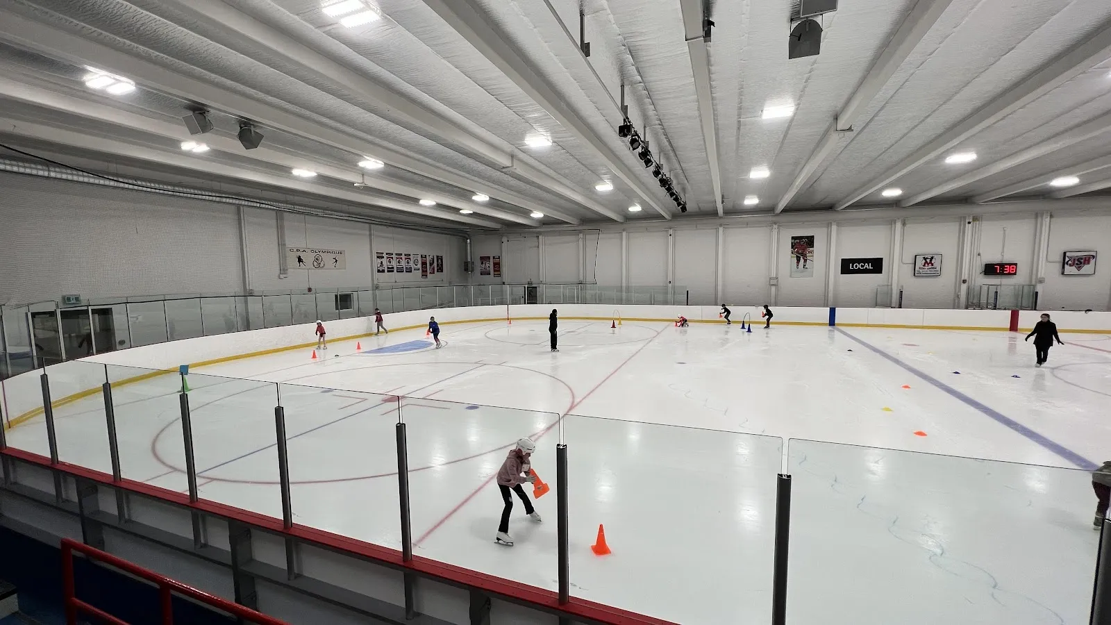 Vue large en hauteur de la surface de glace complète de l’Aréna Francis-Bouillon, avec la rampe d’éclairage, le plafond isolé, les lignes rouge et bleues, les bandes et les baies vitrées, et quelques patineurs répartis sur la glace parmi des cônes d’entraînement. — Aréna Francis-Bouillon