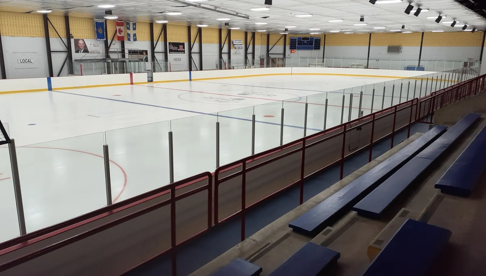 The sheet seen from the bench bleachers, with banners on a yellow wall at the far end and blue lines on fresh ice. — Aréna Gaétan-Duchesne