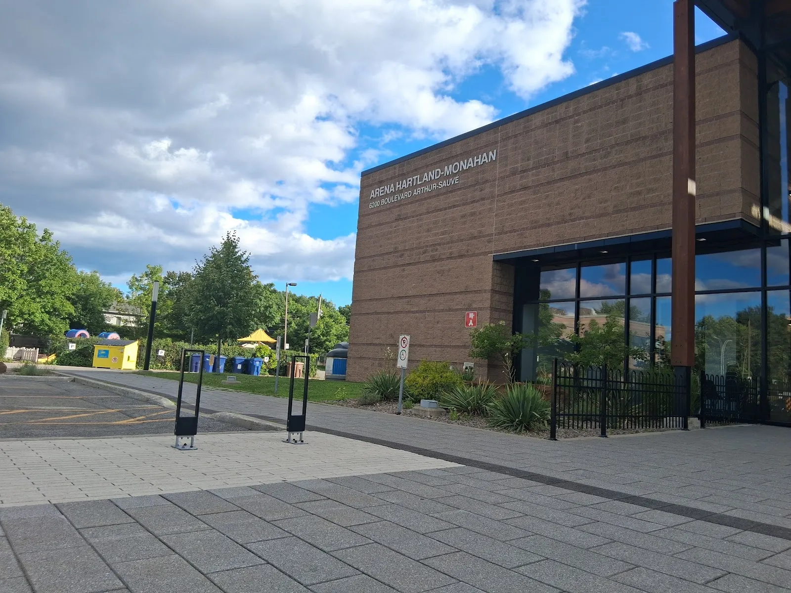 The brick entrance block of Aréna Hartland-Monahan with its name on the wall above tall glass doors, a paved forecourt and young trees under a blue sky. — Aréna Hartland-Monahan (Laval)