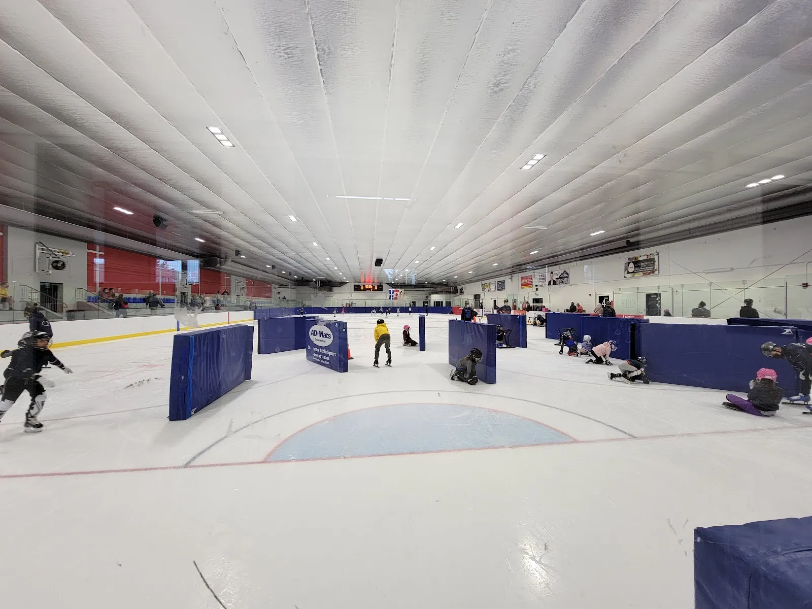 The ice sheet during a learn-to-skate session, seen from the boards: padded blue dividers across the ice, small skaters between them and the white insulated roof above. — Aréna Hartland-Monahan (Laval)