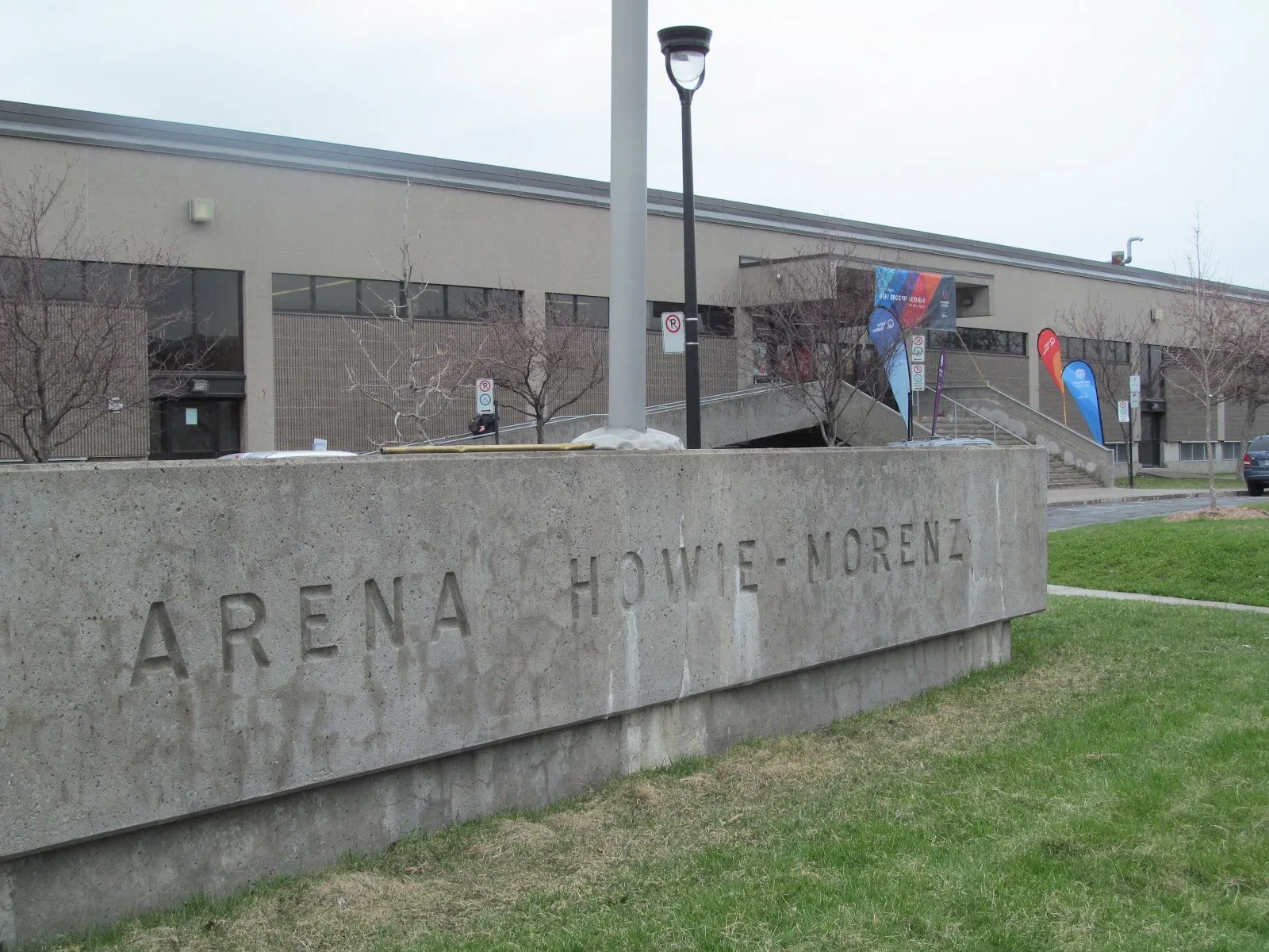 El muro tallado de Arena Howie-Morenz sobre el pasto, con el largo y bajo edificio de la arena y la escalera de su entrada detrás — Aréna Howie-Morenz