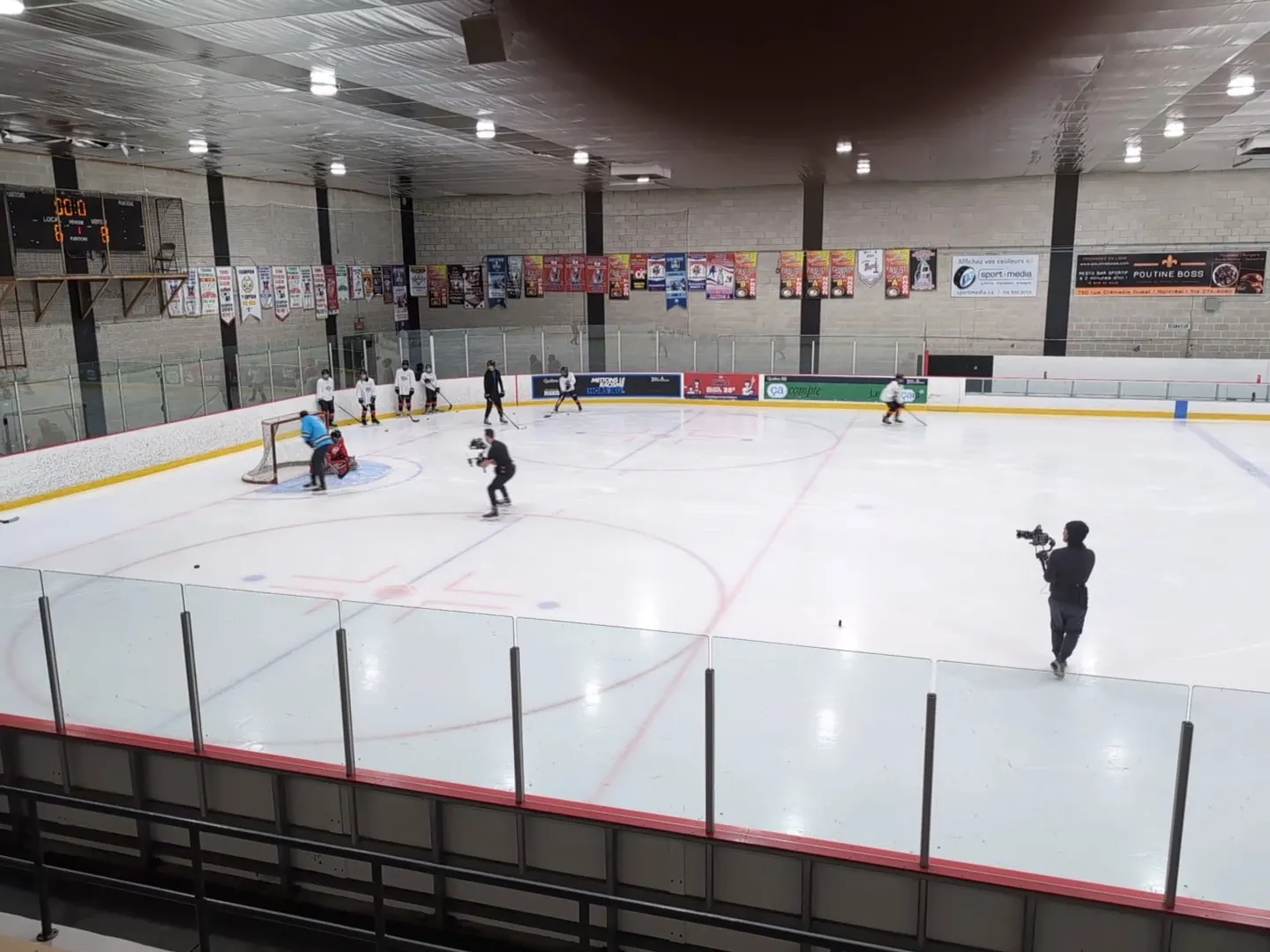 Un partido de hockey sobre trineo en una pista amplia visto por encima del vidrio, con jugadores repartidos cerca de los círculos de saque bajo un techo oscuro con vigas de acero. — Aréna Howie-Morenz