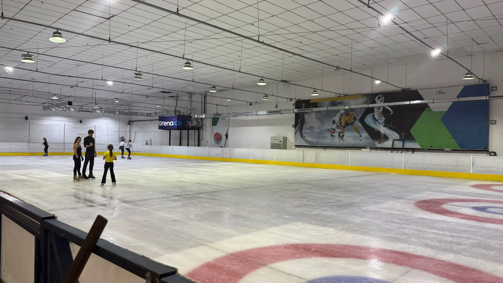 A ampla superfície de gelo da Arena Ice Brasil, com a placa azul da Arena Ice, um círculo de curling em primeiro plano e alguns patinadores ao fundo — Arena Ice Brasil