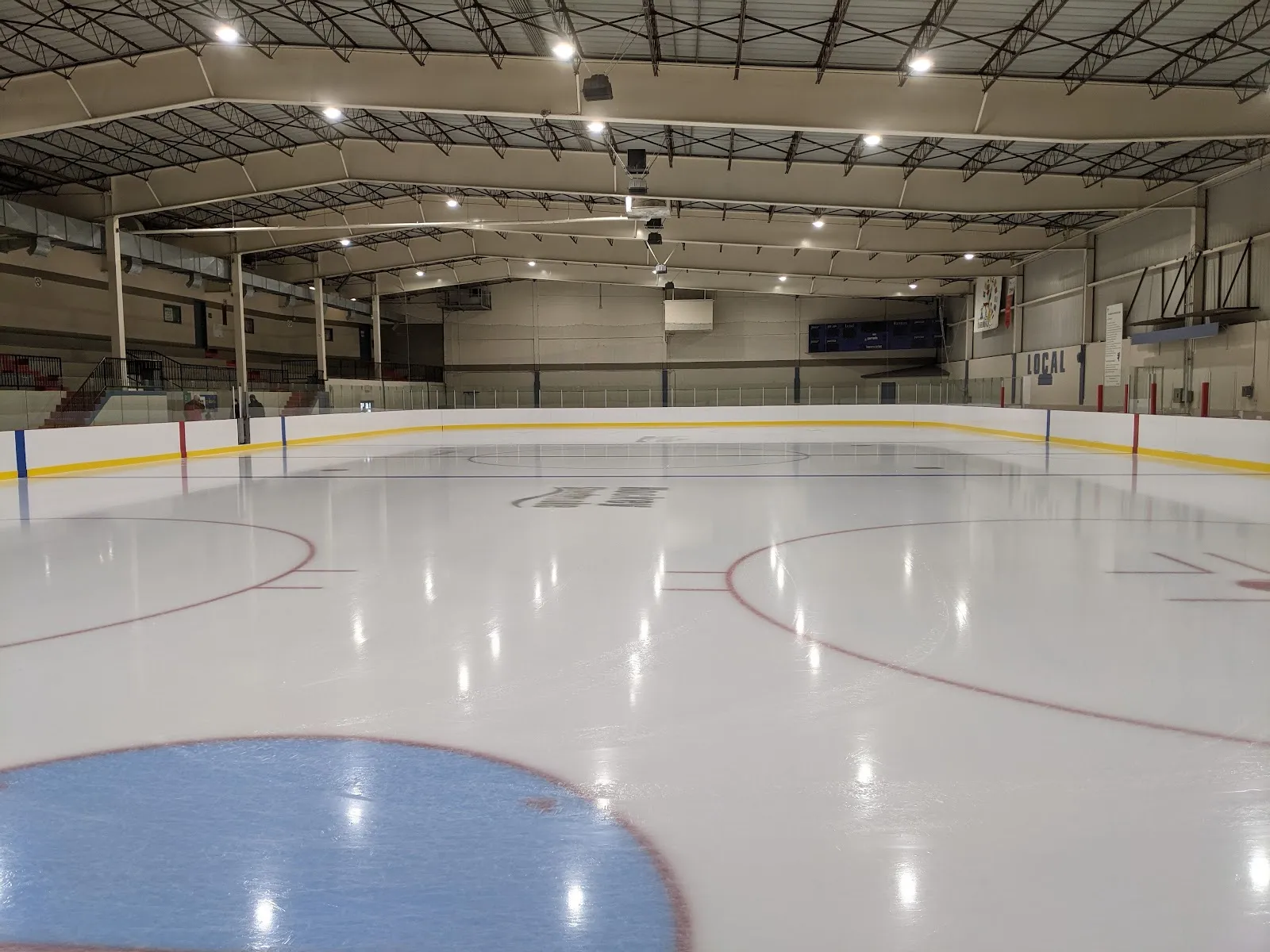 La patinoire vide vue au ras de la glace près d’un cercle de mise au jeu, sous un toit d’acier voûté, avec quelques rangées de gradins d’un côté. — Aréna Ivan-Dugré