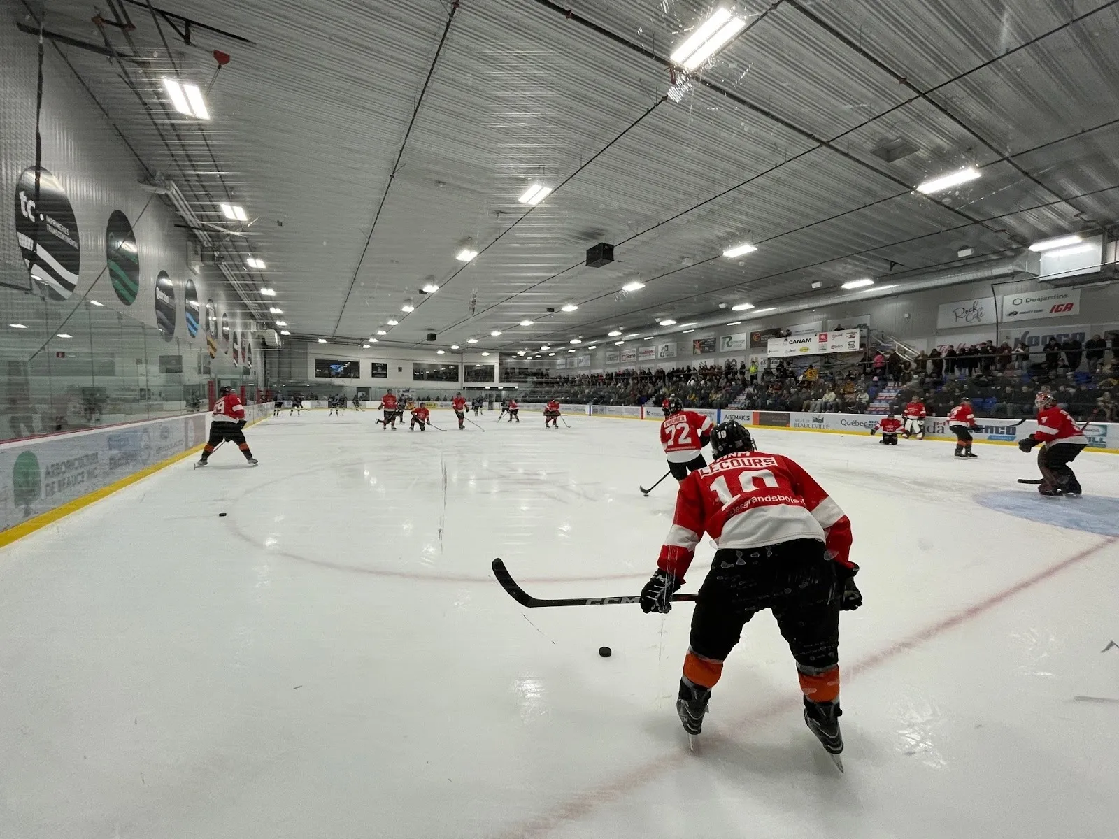 Vue d’ensemble d’un match de hockey depuis la bande, avec des spectateurs dans les gradins du côté opposé et un joueur en chandail rouge au premier plan. — Aréna J.E. Métivier