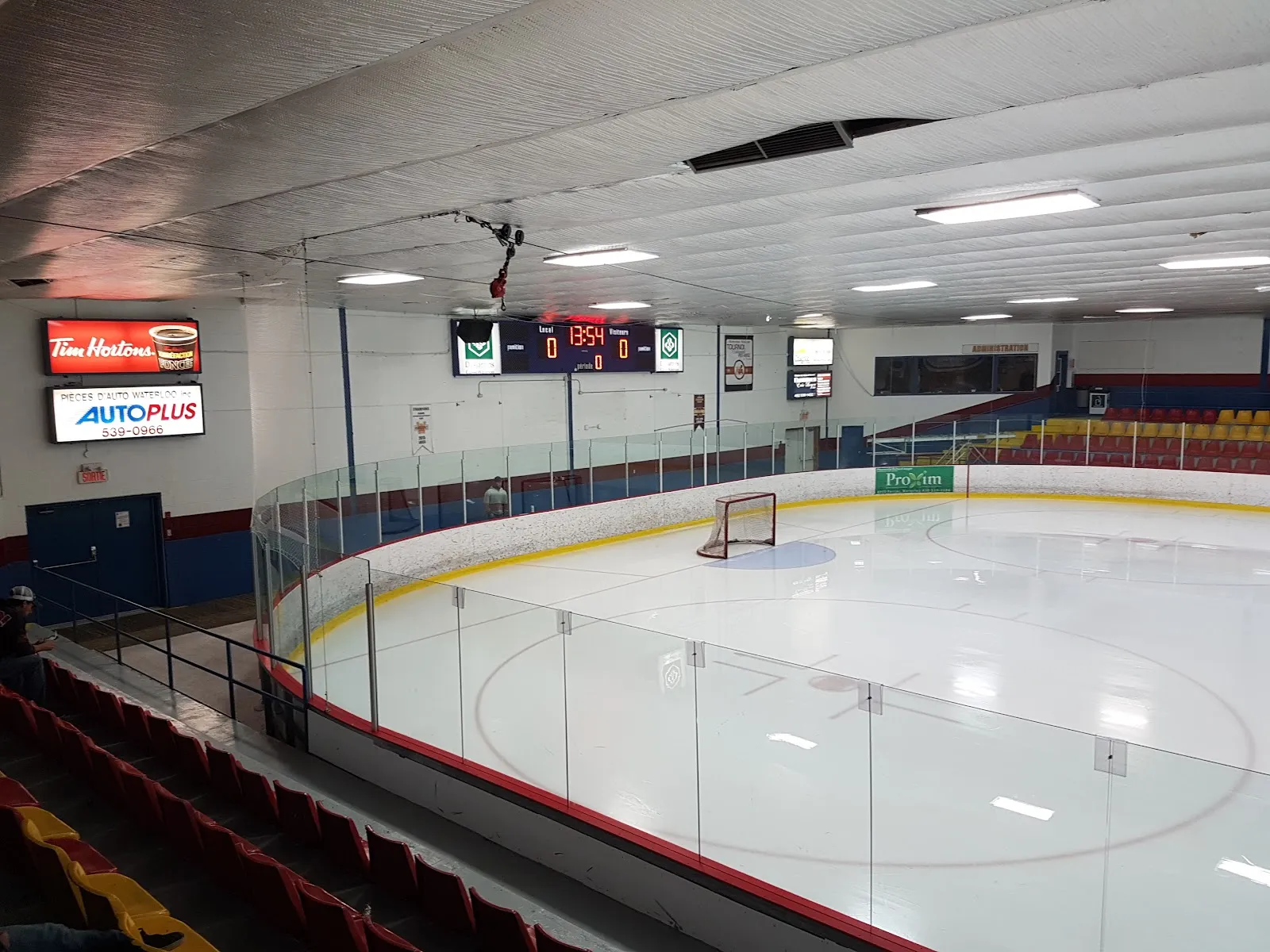 The ice of the Aréna Jacques-Chagnon seen from the stands, with the curved glass, the scoreboard and the red and blue boards — Aréna Jacques-Chagnon