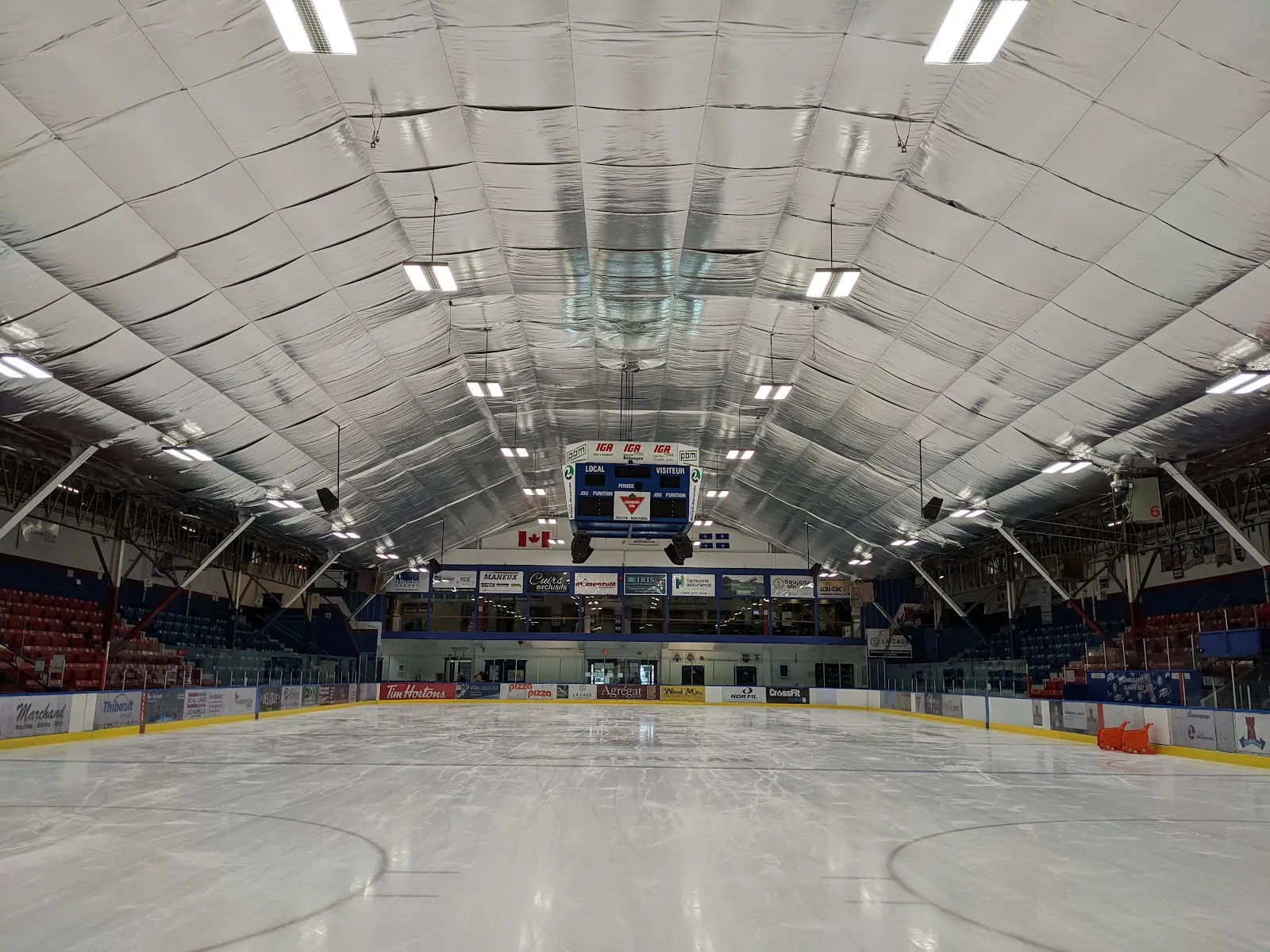 The empty ice of the Aréna Jacques-Laperrière seen from the end of the rink, with the red and blue seating, the scoreboard and the arched ceiling. — Aréna Jacques-Laperrière