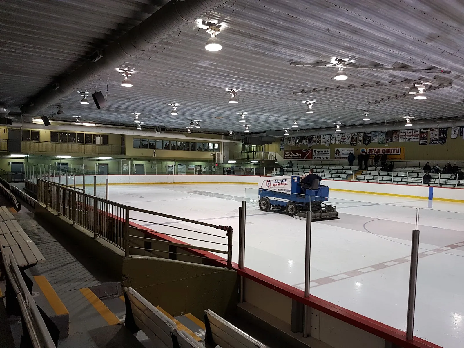 Une surfaceuse traversant la patinoire, vue des bancs du fond, avec l’étage vitré le long du côté. — Aréna Jean-Rougeau
