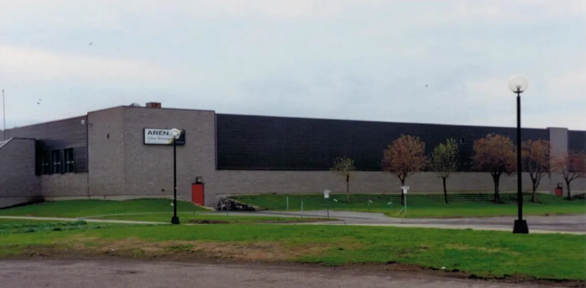 The long brick and dark-panel wall of the Aréna Julien-Beauregard in Marieville, with its arena sign on the corner block, seen across the lawn — Aréna Julien-Beauregard