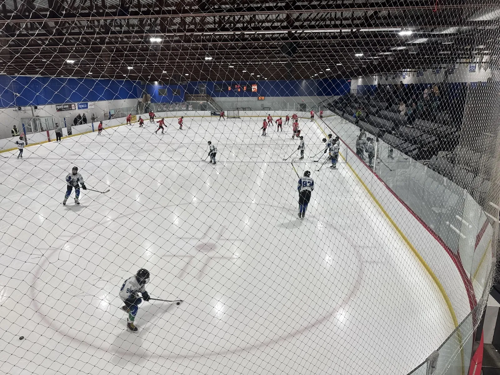 A game under a dark steel ceiling seen through netting, players spread across the white ice with a blue board band behind. — Aréna Julien-Beauregard