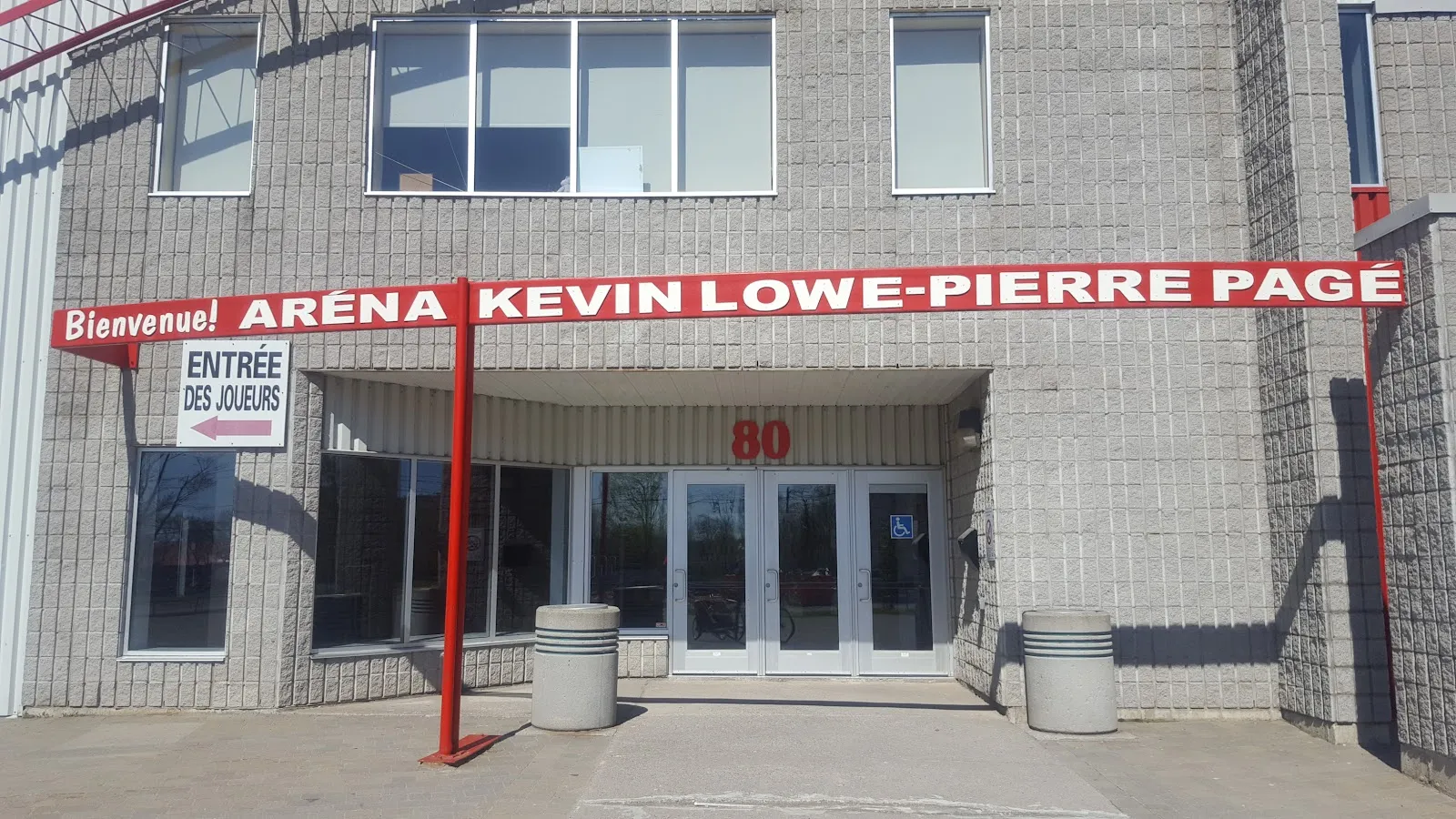 The brick entrance of the Aréna Kevin-Lowe–Pierre-Pagé under its red welcome sign, number 80 above the doors — Aréna Kevin-Lowe–Pierre-Pagé