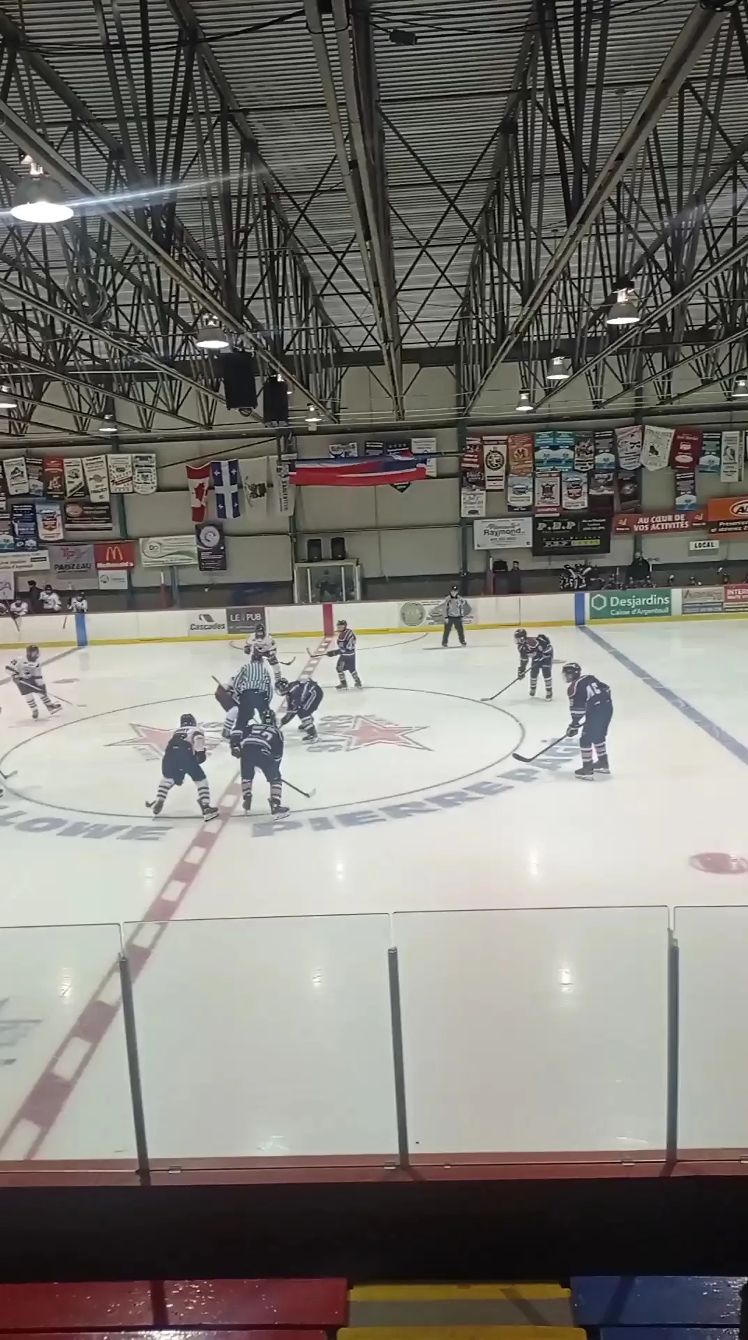 A hockey game under way at the Aréna Kevin-Lowe–Pierre-Pagé, with the steel roof trusses and sponsor banners — Aréna Kevin-Lowe–Pierre-Pagé