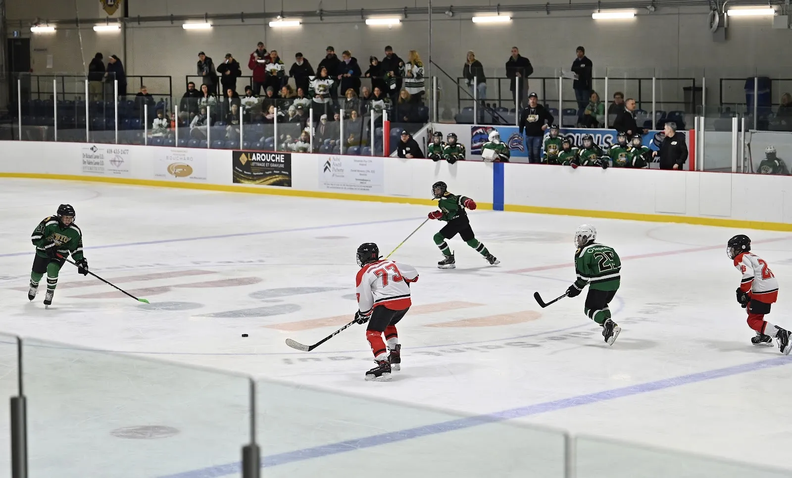 A game under a dark ceiling at the far end of a wide sheet, players in green and white spread across the ice below the stands. — Aréna Luc-et-Marie-Claude