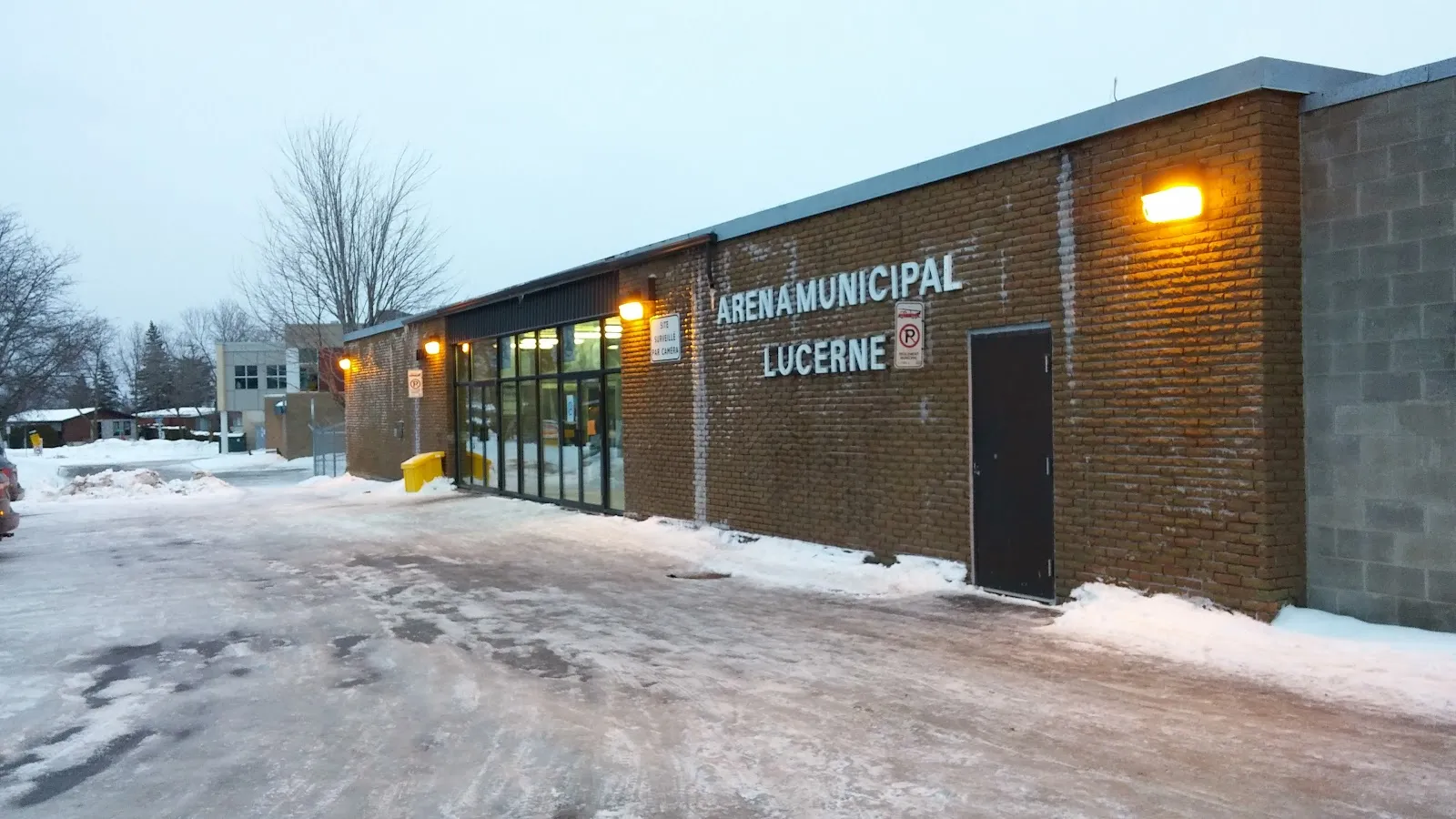 Le long bâtiment en brique de l'Aréna municipal Lucerne avec son nom en lettres blanches, l'entrée vitrée et une porte brune, par un soir de neige avec les lampes murales allumées. — Aréna Lucerne (Laval)