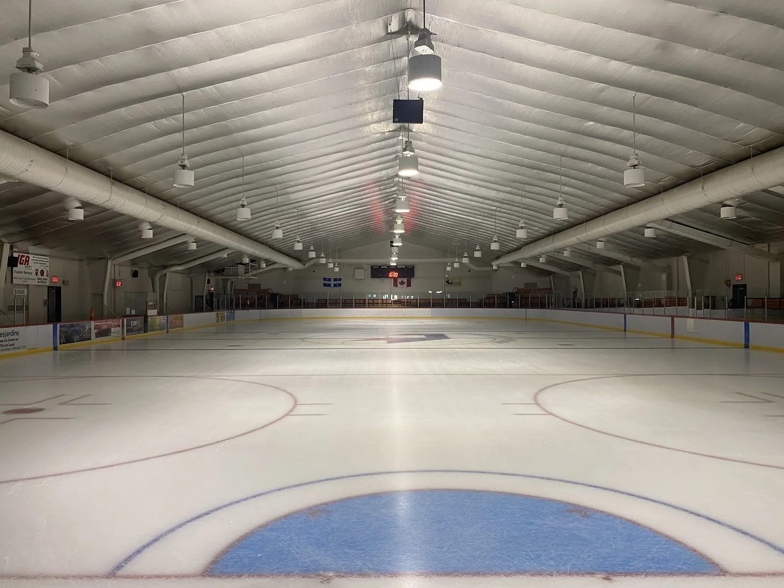 La surface de glace vide vue depuis la bande, avec le cercle de mise au jeu bleu au premier plan, le long toit isolé, les luminaires suspendus et la bande du fond. — Aréna Lucerne (Laval)