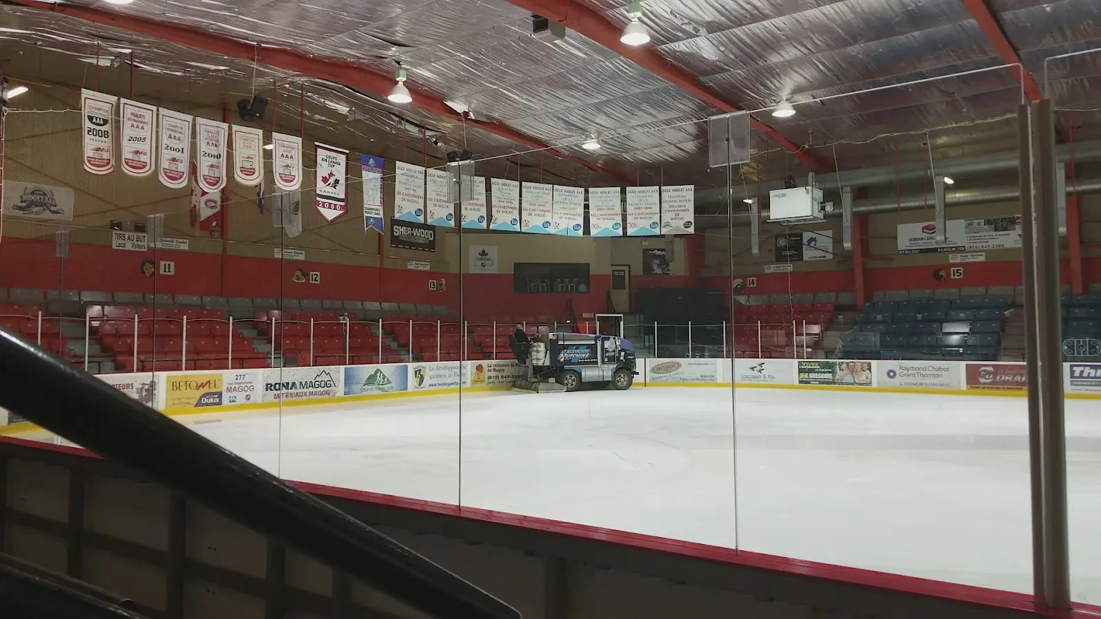 The ice sheet at the Aréna de Magog with the resurfacer at the far end, banners hanging above the boards and red seating behind the glass. — Aréna de Magog