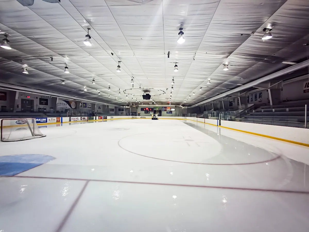 La patinoire vide vue au ras de la glace à une extrémité, sous un plafond isolé blanc garni de rangées de lumières, avec des gradins le long du côté droit. — Aréna Marcel-Larocque