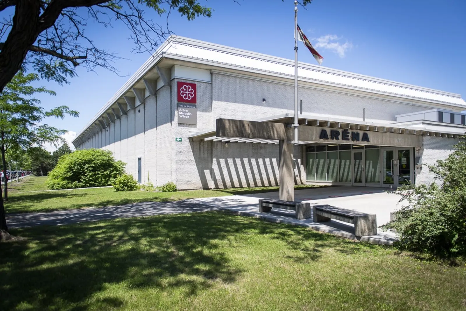Entrada principal de Aréna Marcelin-Wilson en un día soleado: un edificio bajo de ladrillo blanco con una placa roja de la Ville de Montréal y un letrero de "Aréna Marcelin-Wilson", un asta de bandera y una marquesina techada de concreto que dice "ARÉNA" sobre las puertas de vidrio. — Aréna Marcelin-Wilson