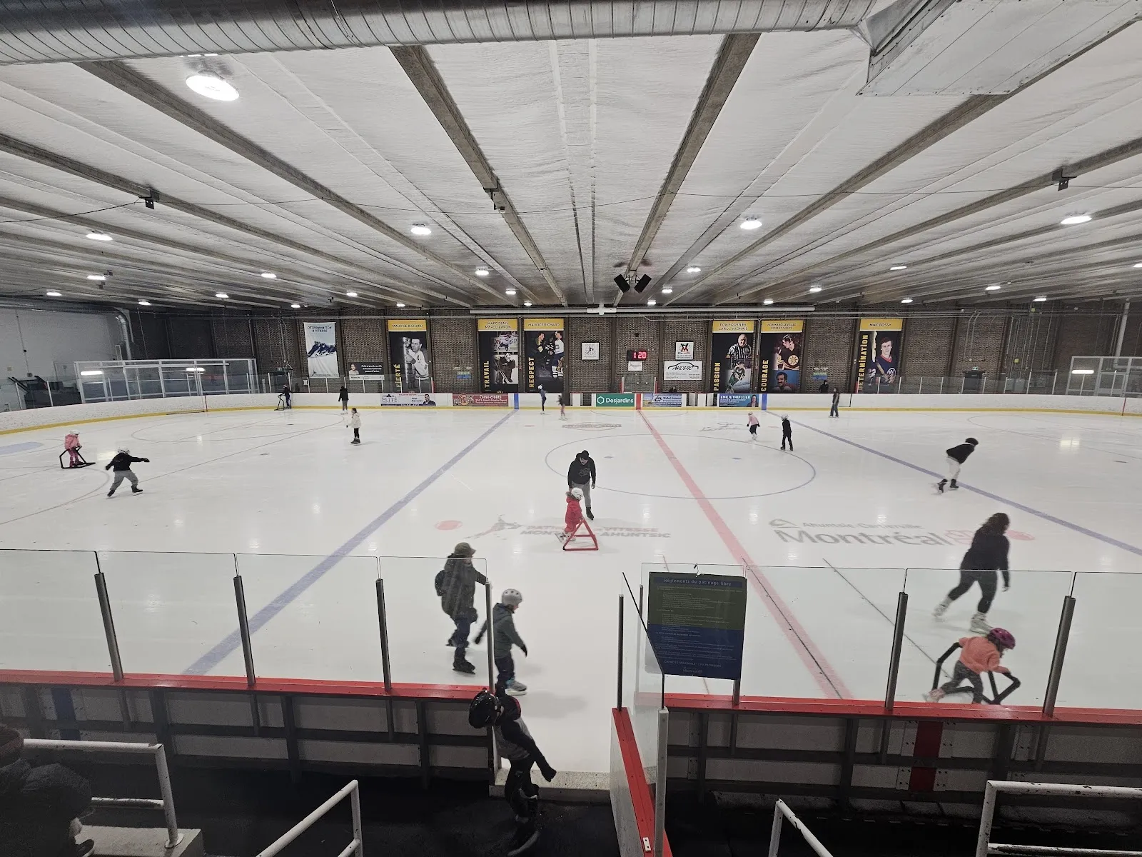 Una sesión pública repartida por una pista amplia, con patinadores y algunas sombras de jugadores en el hielo bajo un techo blanco acanalado. — Aréna Marcelin-Wilson