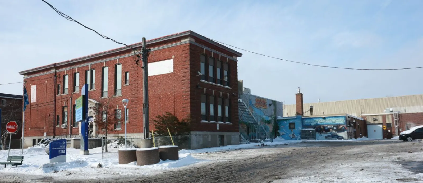 Corner view of the old two-storey red-brick arena building in winter, with a blue "ARÉNA MARTIN-LAPOINTE" wayfinding sign and municipal parking sign in the foreground and a hockey-themed mural visible further along the wall. — Aréna Martin-Lapointe