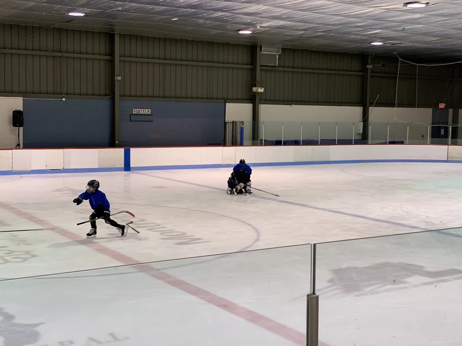 A wide sheet with two young skaters on it, red and blue lines on pale ice and a steel-framed roof over a blue-trimmed far wall. — Aréna Martin-Lapointe