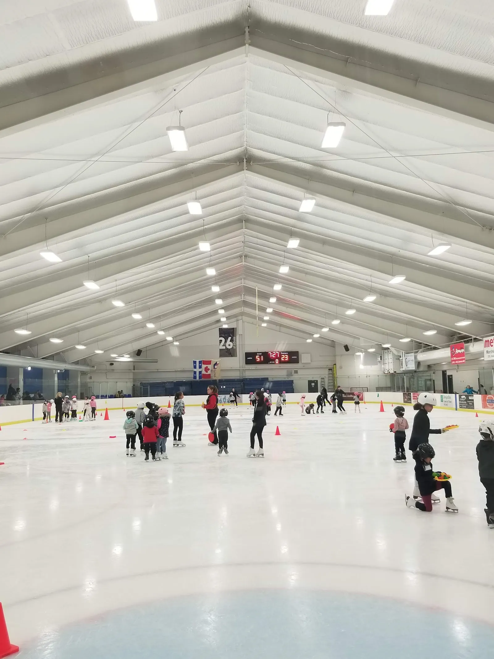 The whole ice sheet from the boards during a children's skating class, with small skaters and orange cones across the ice, the Québec and Canadian flags, the scoreboard and a retired-number banner under the white vaulted roof. — Aréna Martin-St-Louis (Laval)